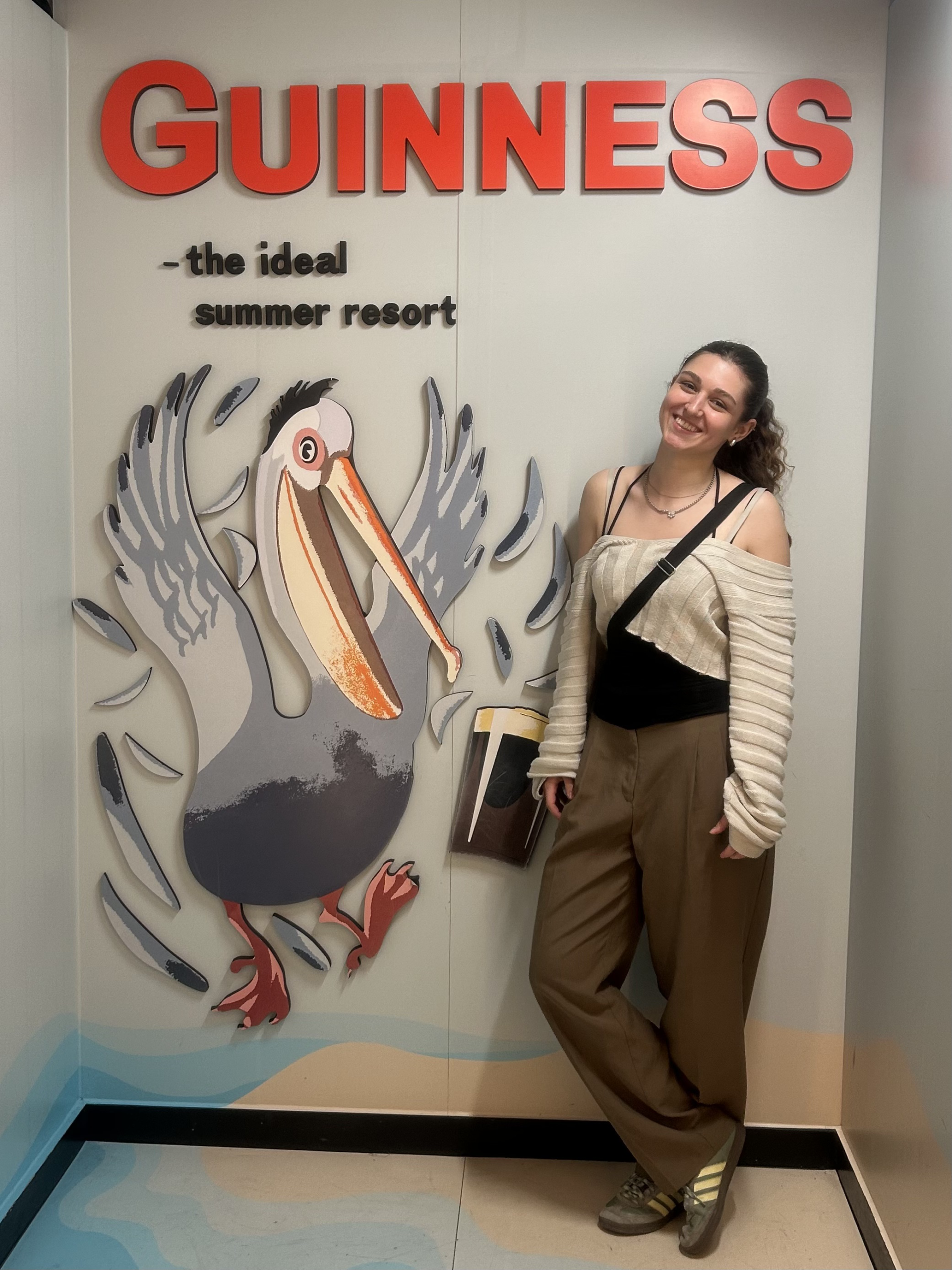 Leonie steht vor einem Guiness Schild mit der Aufschrift: "the ideal summer resort". Unter der Aufschrift ist ein gezeichneter Pelikan und ein Glas Guiness. Leoni lehnt daneben an der Wand. Sie trägt ein weißes, weites Crop Top, ein Schwarzes Trägertop und eine weite grün-graue Hose.